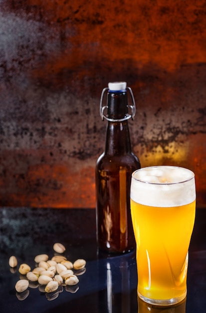 Glass with freshly poured unfiltered light beer, beer bottle near scattered pistachios on a black mirror surface. Food and beverages concept