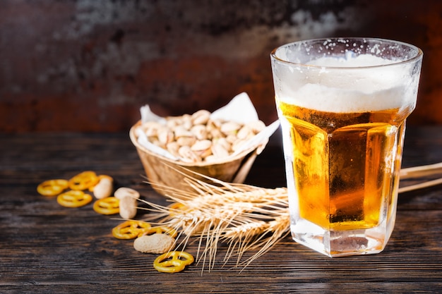 Glass with light beer and a head of foam near plate with pistachios, wheat, scattered small pretzels and peanuts on dark desk. Food and beverages concept