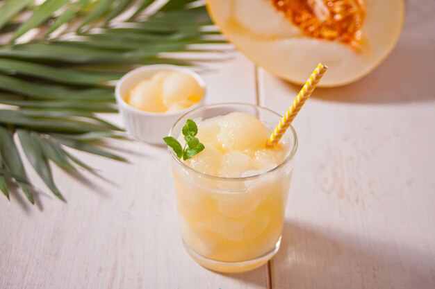 Glass with melon cocktail on a white table with the palm leaf in tropical theme