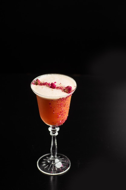 Glass with orange cocktail decorated with dried rose on the bar on a black background