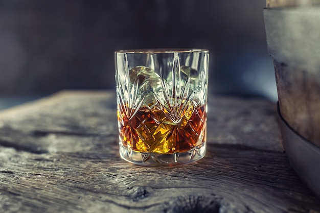 Glass with whiskey rum brandy or bourbon on an old table near a wooden barrel.