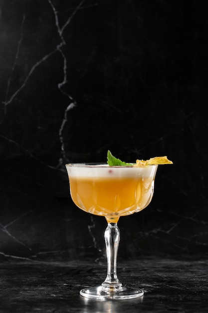 Glass with yellow lemon cocktail decorated with dried pineapple and mint on the bar on a black