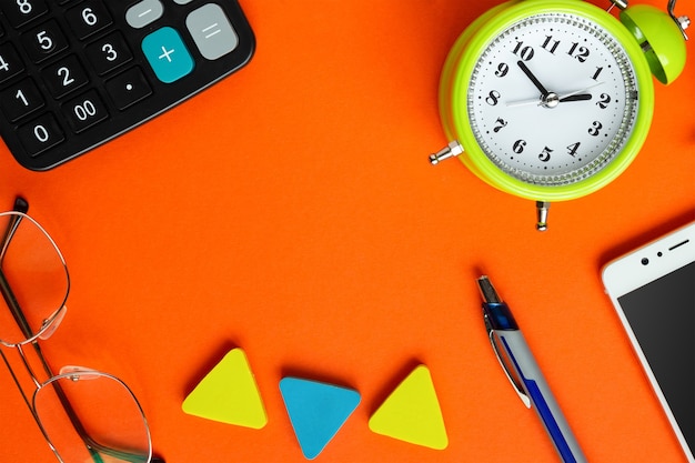 Glasses, alarm clock, smartphone, calculator and pen on a bright orange table.