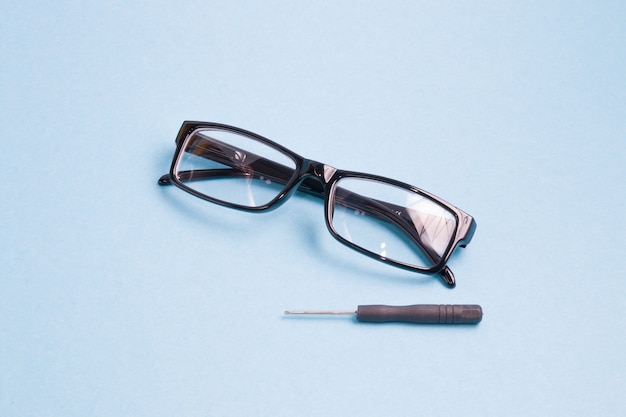 Glasses in a black plastic frame and a small screwdriver are on a light blue surface