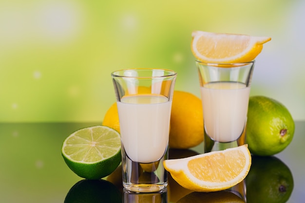 Glasses of cream liqueur with lime and lemon on green background with reflection. Short of lemon liqueur. Traditional Italian alcoholic beverage Limoncello