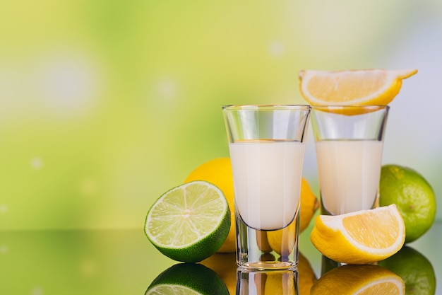 Glasses of cream liqueur with lime and lemon on green background with reflection. Short of lemon liqueur. Traditional Italian alcoholic beverage Limoncello