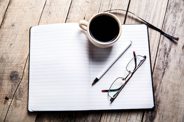 Glasses on notebook with pen and cup of coffee in wood table