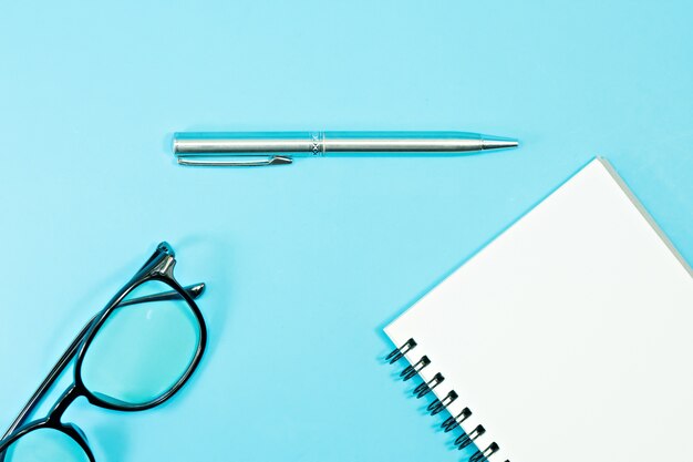 Glasses and pen with blank notebook on blue background