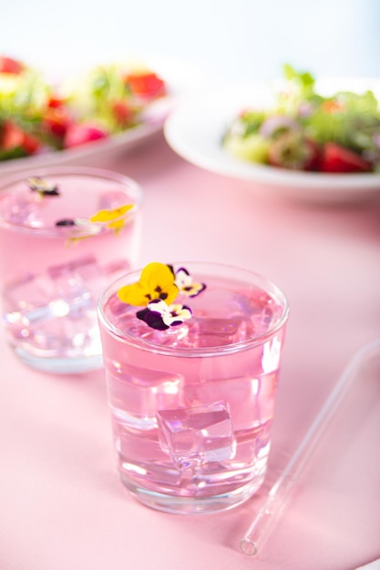 Glasses of pink violet purple cocktail decorated flowers viola pansy and fresh salad on the background