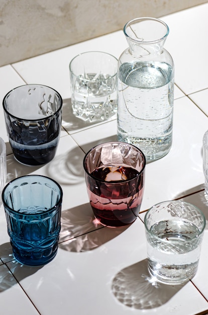 Glasses of water from multi-colored glass on white tiles, hard\
rays of the sun. quench your thirst, drink water