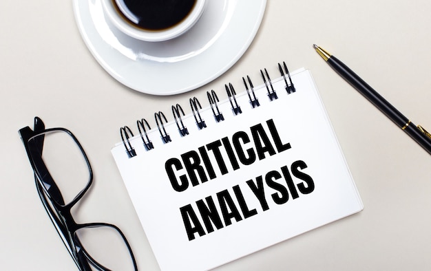 眼鏡、白い一杯のコーヒー、CRITICAL ANALYSISという言葉が書かれた白いノート、ボールペンが明るい面に横たわっています。