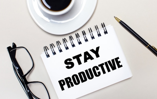 Очки, белая чашка кофе, белый блокнот с надписью STAY PRODUCTIVE и шариковая ручка лежат на светлом фоне.