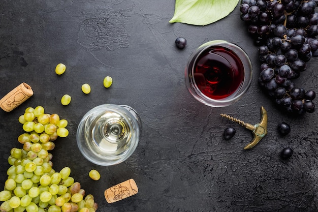 Glasses of white and red wine with ripe grapes on black stone background, top view
