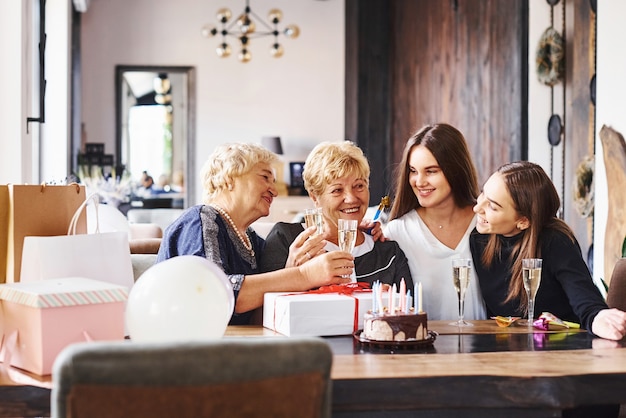 Glasses with alcohol in hands and cake on table. Senior woman with family and friends celebrating a birthday indoors.