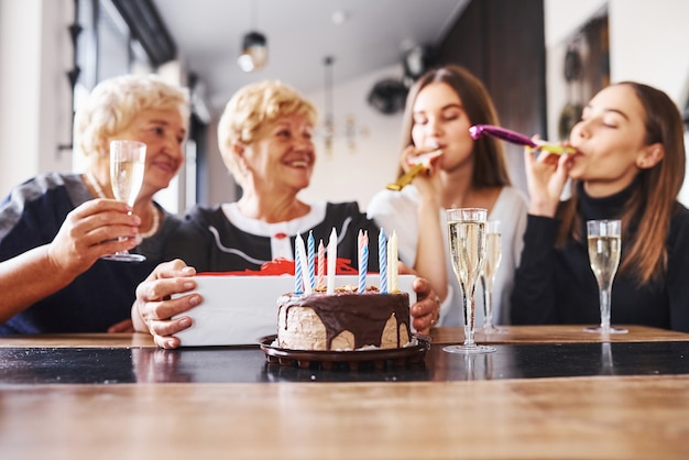 手にアルコールとテーブルの上のケーキとグラス。屋内で誕生日を祝う家族や友人と年配の女性。