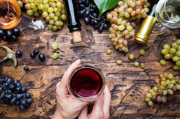 Glasses with white, red and pink wine and ripe grapes on wooden background, top view. womans hand with wine glass