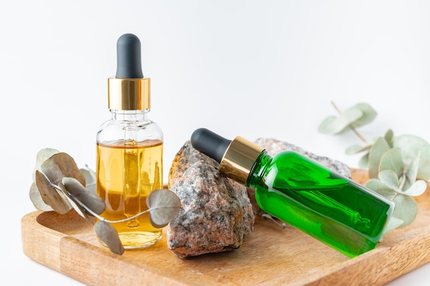 Glazen druppelflesjes met een pippette met gouden rubberen tip en groene eucalyptus bladeren in de buurt van het op houten dienblad op witte achtergrond Nature Skin concept Organic Spa Cosmetics