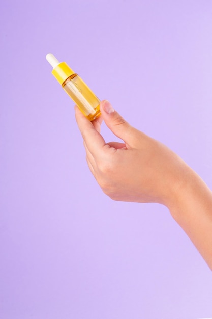 glazen gele fles met pipet met etherische olie in vrouwelijke hand op paarse achtergrond bovenaanzicht.