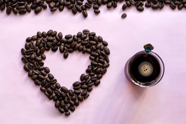 Glazen koffiemok en gebrande koffiebonen op een roze achtergrond. Koffiemanie, liefde voor koffie, concept. Hart van bonen. bovenaanzicht
