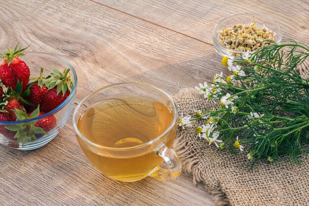 Glazen kopje groene thee, kamille bloemen op zak, glazen kommen met droge bloemen van matricaria chamomilla en verse aardbeien op de houten planken.