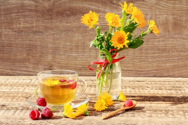 Glazen kopje groene thee met bloemen van calendula en verse frambozen, vers boeket van oranje calendula in vaas. Houten planken achtergrond. Gezondheidsdrankje.