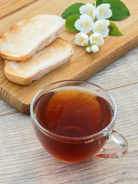 Glazen kopje thee, brood en witte jasmijn bloemen op houten snijplank. Bovenaanzicht.