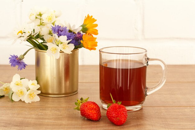 Glazen kopje thee met aardbeien en bloemen op houten bureaublad.