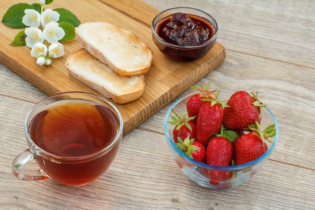 Glazen kopje thee, zelfgemaakte aardbeienjam, brood op houten snijplank en verse aardbeien, witte jasmijnbloemen op houten achtergrond. Bovenaanzicht.