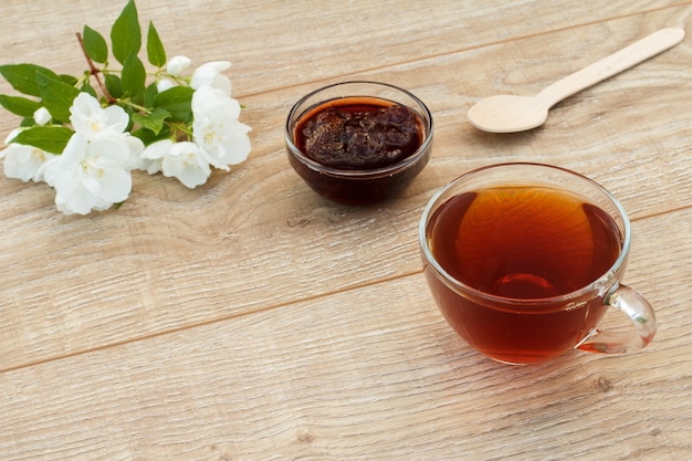 Glazen kopje thee, zelfgemaakte aardbeienjam, houten lepel en witte jasmijn bloemen op houten achtergrond. bovenaanzicht.