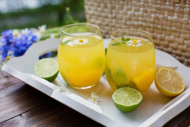 Glazen met limonade met muntlimoen, citroenen en sinaasappel op een wit houten dienblad in de buurt van strozak met wilde bloemen Zomerdrankenconcept