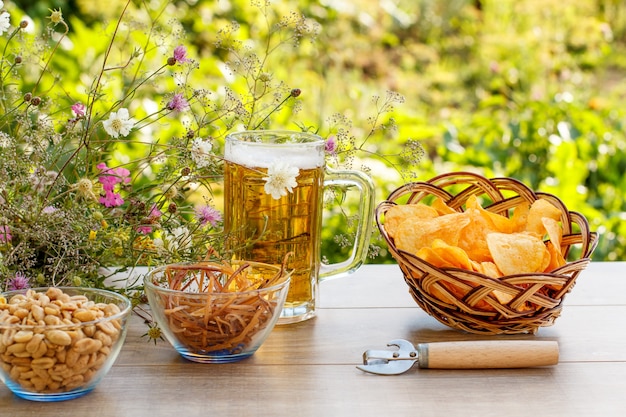 Glazen mok bier op houten tafel met chips in rieten mand, pinda's en gedroogde inktvis in kommen op natuurlijke groene onscherpe achtergrond