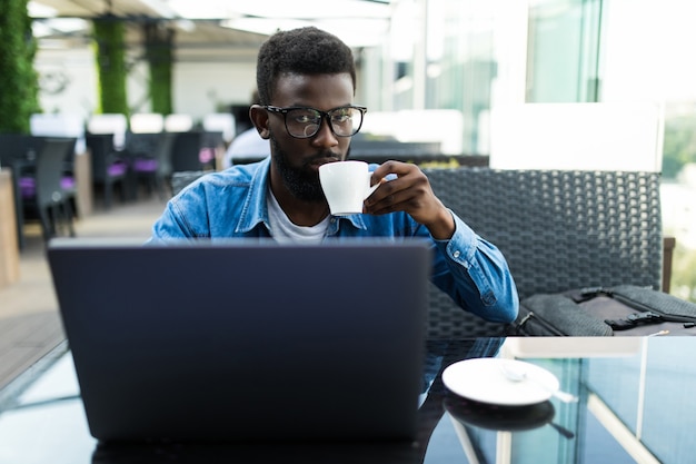 Foto glimlach afrikaanse zakenman zittend in café met een kopje koffie en met behulp van laptop.