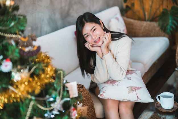 Glimlach geluk portret mooie jonge aziatische vrouw die warme kleren draagt, zittend op de bank met koffiekopje mok decoratie tijdens kerst, kerstmis en nieuwjaarsvakanties thuis in de kamer binnen of in het café