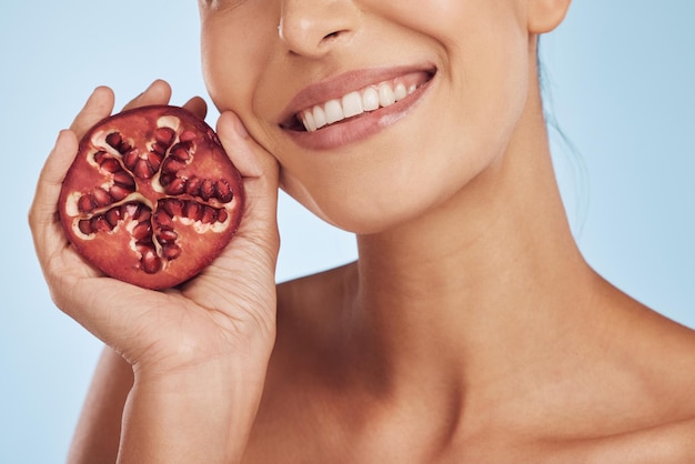 Glimlach huidverzorging en handen van vrouw met granaatappel in studio geïsoleerd op een blauwe achtergrond Natuurlijk fruit en gezicht van model met voedsel voor voeding gezond veganistisch dieet en vitamine c voor wellness
