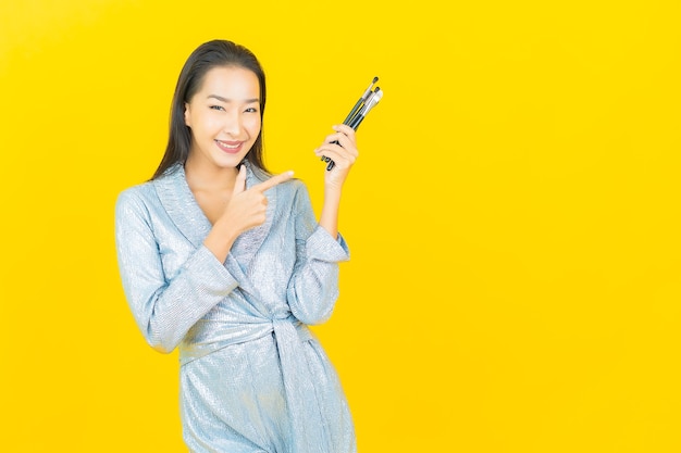 Glimlach van de portret maakt de mooie jonge aziatische vrouw met schoonheidsmiddel omhoog borstel op gele muur