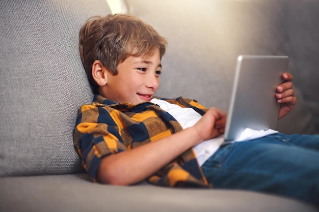 Glimlachen bank en jongen kind met tablet online spel en zoeken naar video streaming op de website Scroll ontspannen en gelukkig kind op de bank met digitale app voor elearning internetverbinding en plezier thuis