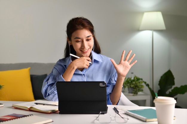 Glimlachen en hallo zeggen. Gelukkige Aziatische vrouw die oortelefoons draagt met een videogesprek op een tablet die thuis op een bureau in de woonkamer zit. Jonge Aziatische vrouw entrepreneu die online educatieve lessen geeft