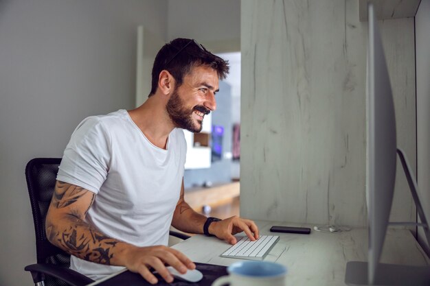 Glimlachend aantrekkelijke bebaarde getatoeëerde freelancer om thuiskantoor te zitten en computer te gebruiken.