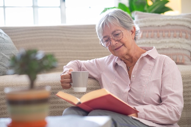 Glimlachend aantrekkelijke senior vrouw ontspannen zittend op de vloer een boek lezen met een kopje koffie of thee Kaukasische volwassen dame studeren leren thuis