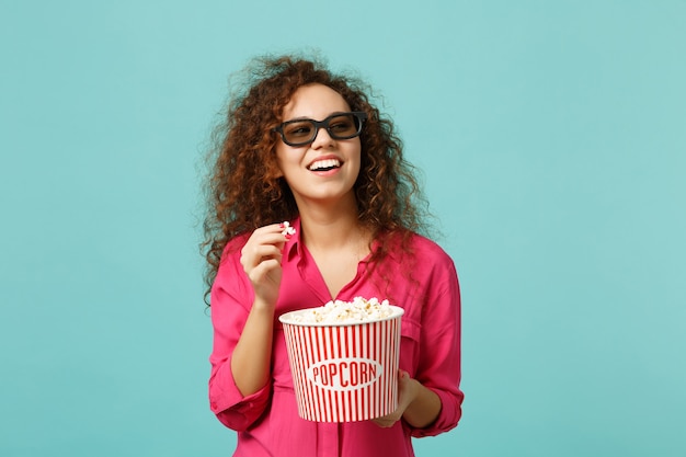 Glimlachend afrikaans meisje in 3d imax bril kijken naar film film en houden popcorn geïsoleerd op blauwe turquoise muur achtergrond in studio. mensen emoties in de bioscoop, lifestyle concept. bespotten kopie ruimte.