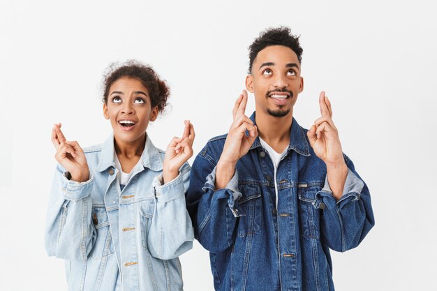 Glimlachend Afrikaans paar in denimoverhemden die samen met gekruiste vingers bidden terwijl omhoog het kijken over grijze muur