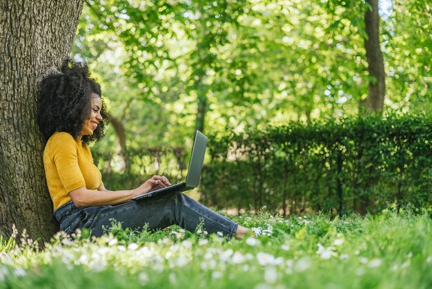 Glimlachend afro meisje chatten met een laptop in een park zittend tegen een boom