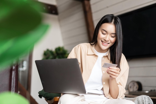 Glimlachend Aziatisch meisje dat betaalt voor online winkelen op laptop en naar haar creditcard kijkt om thuis te zitten...