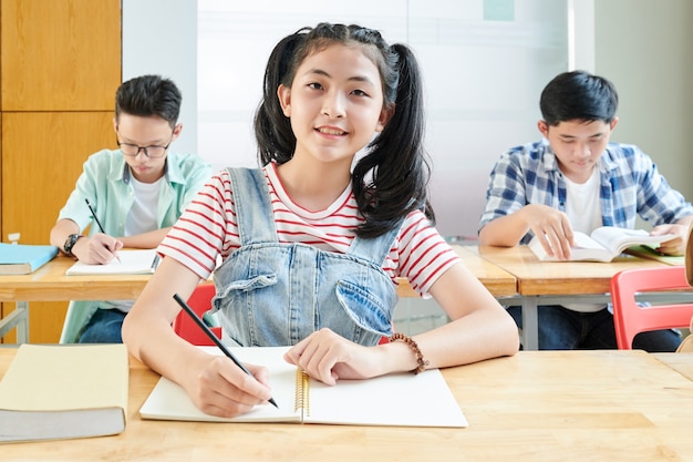 Glimlachend Aziatisch schoolmeisje dat essay in leerboek schrijft tijdens Engelse les