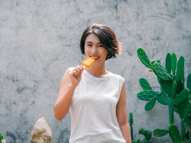 Glimlachend Aziatische vrouw kort haar in casual wit mouwloos shirt met gele ijslolly op grijze betonnen muur achtergrond in de zomer. Vrouwen die ijslollys eten, vers zomerseizoenconcept