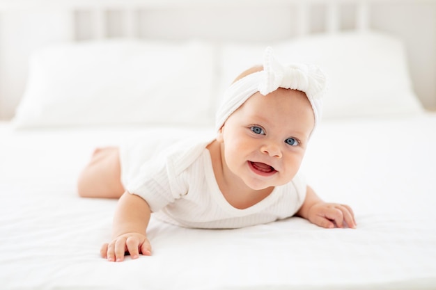 glimlachend babymeisje met blauwe ogen portret close-up gelukkig kleine baby van zes maanden liggend op haar buik in een lichte slaapkamer in een witte bodysuit op het bed en lachen