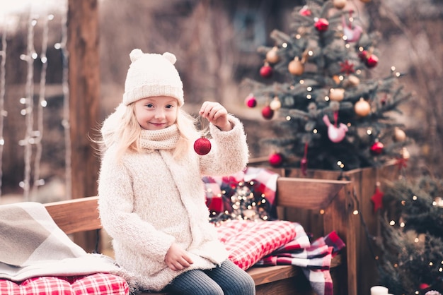 Glimlachend babymeisje met kerstbal zittend op een bankje onder de kerstboom