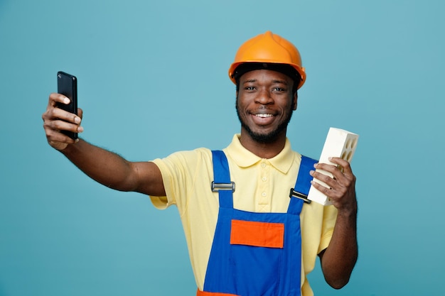 Glimlachend bedrijf baksteen jonge Afro-Amerikaanse bouwer in uniform nemen een selfie geïsoleerd op blauwe achtergrond