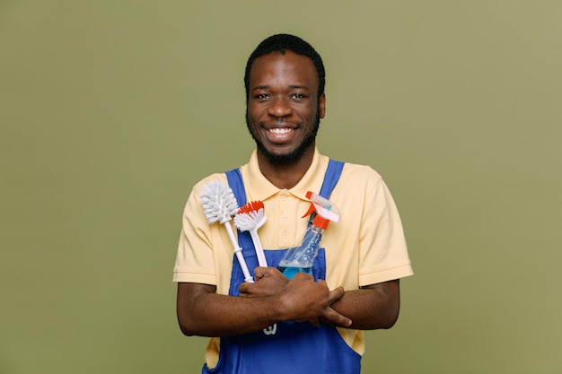Glimlachend bedrijf schoonmaak tools jonge Afro-Amerikaanse schonere man in uniform met handschoenen geïsoleerd op groene achtergrond