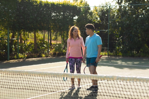 Glimlachend biraciaal koppel dat tennis speelt en praat op de zonnige buitentennisbaan. Inclusiviteit, sport, gezonde hobby's, competitie, fitness en vrije tijd concept.